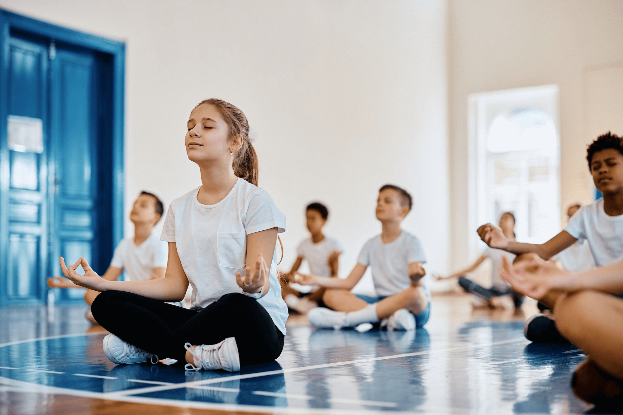 A group of children sit cross-legged on a gym floor with their eyes closed and hands in a meditation pose, wearing casual athletic clothing, in a bright indoor space.