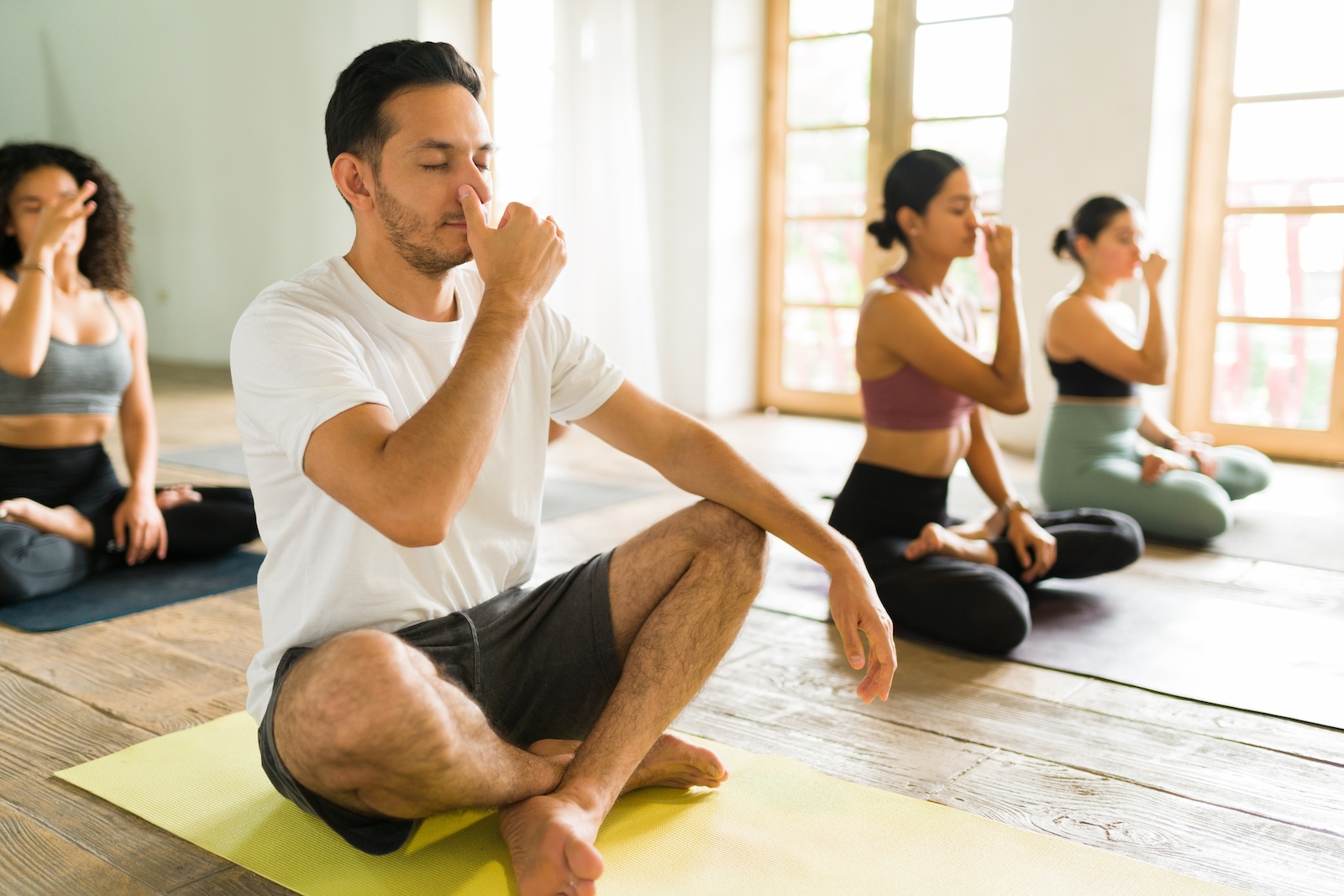 A group of people sitting cross-legged on yoga mats indoors practice alternate nostril breathing. The man in the front pinches his nose with his fingers. Large windows let in sunlight.
