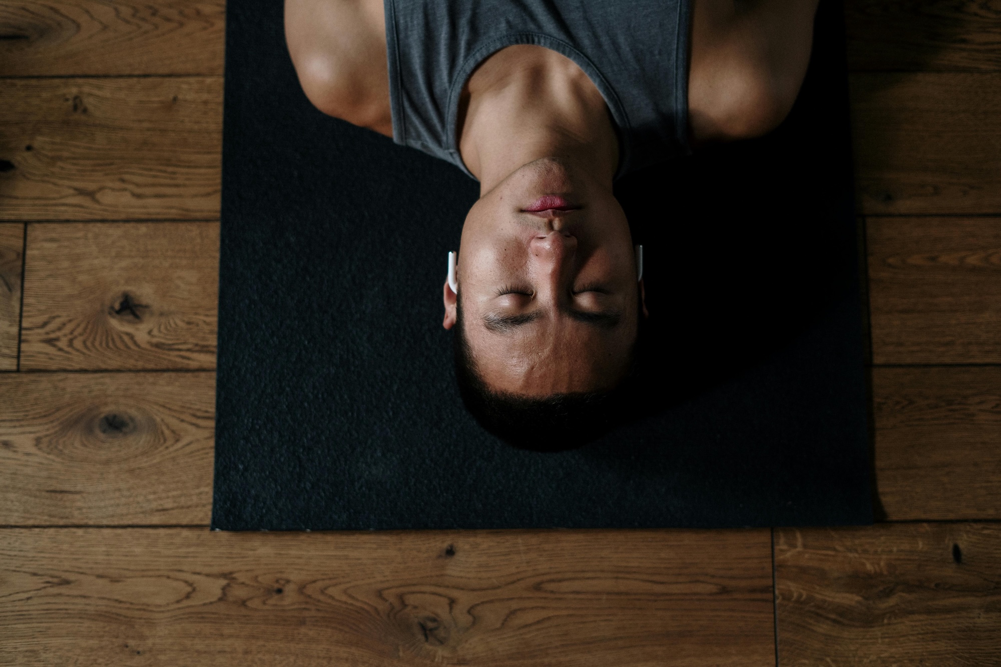 A person wearing a gray tank top and wireless earbuds lies on a black exercise mat with eyes closed on a wooden floor, viewed from above.
