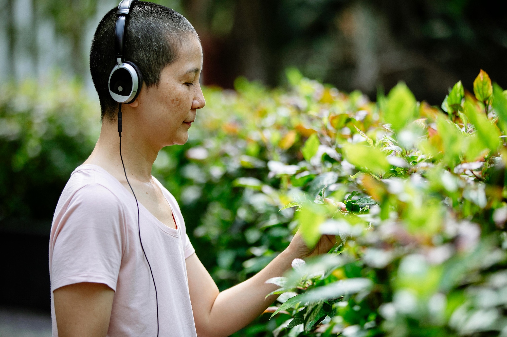 A person with short hair wearing headphones and a light pink shirt touches green leaves on a hedge, standing outdoors surrounded by plants.