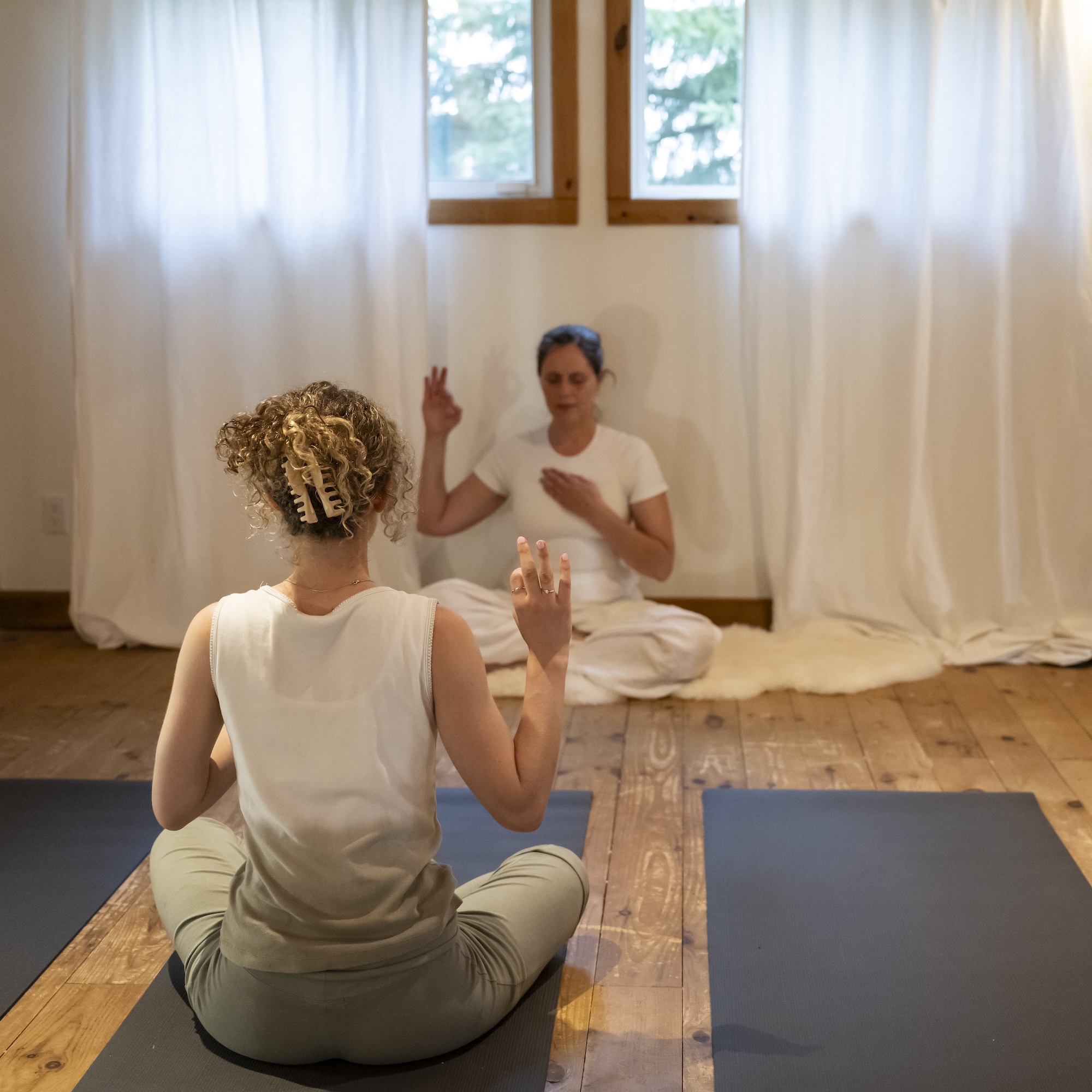 Two people sit on yoga mats in a bright room with wooden floors and white curtains. The person in front sits cross-legged, facing a second person who also sits cross-legged and gestures with one hand raised.