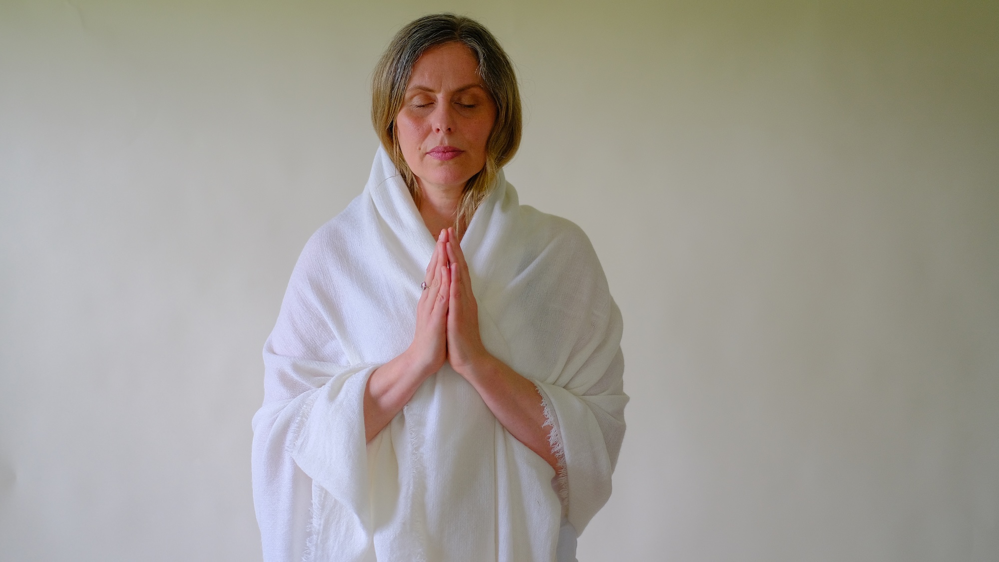 A woman wearing a white shawl stands against a plain background with her eyes closed and hands pressed together in a prayer position.