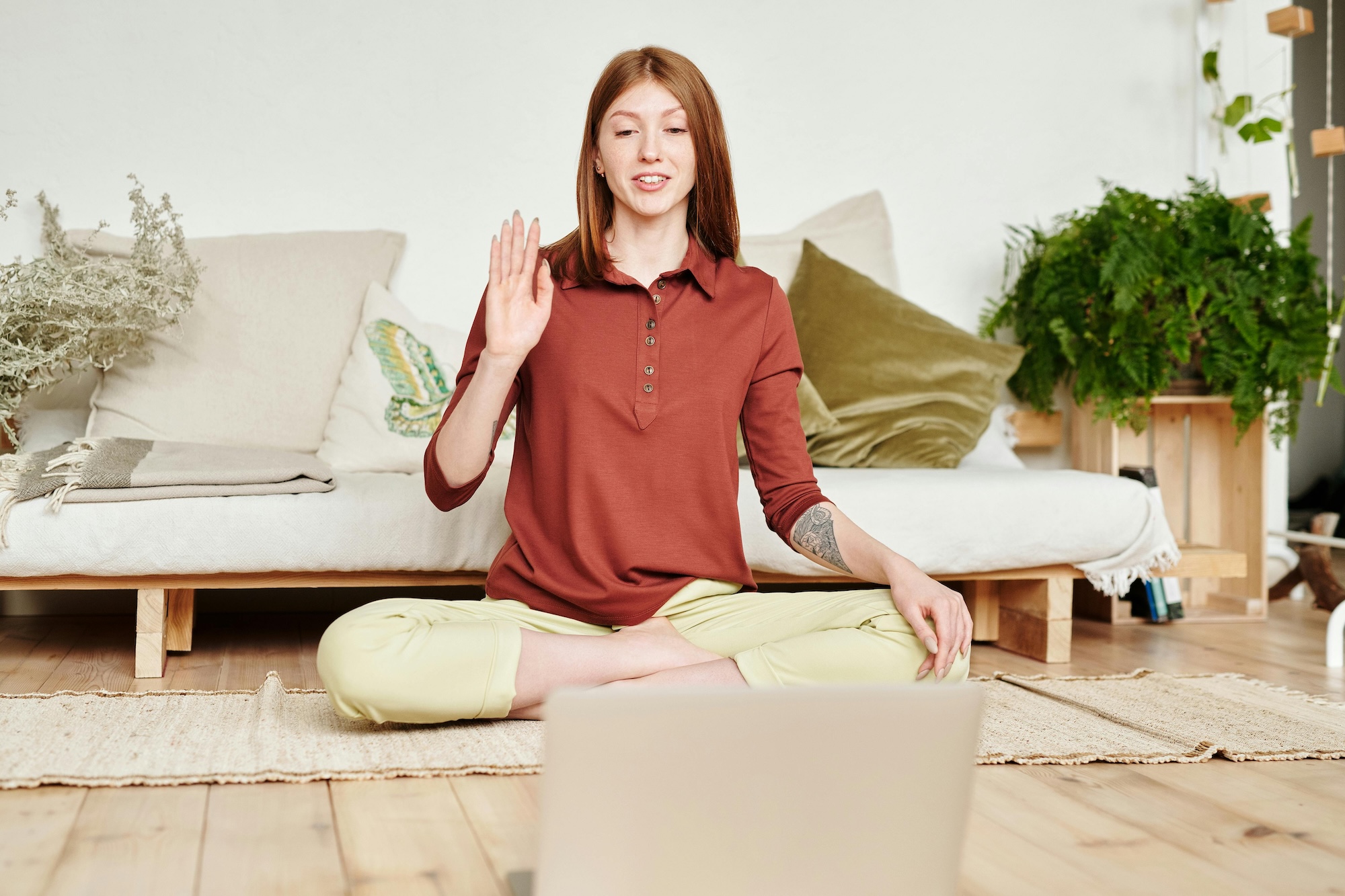 A woman with red hair sits cross-legged on the floor in front of a laptop, waving and smiling, in a bright room with a sofa, plants, and cushions in the background.