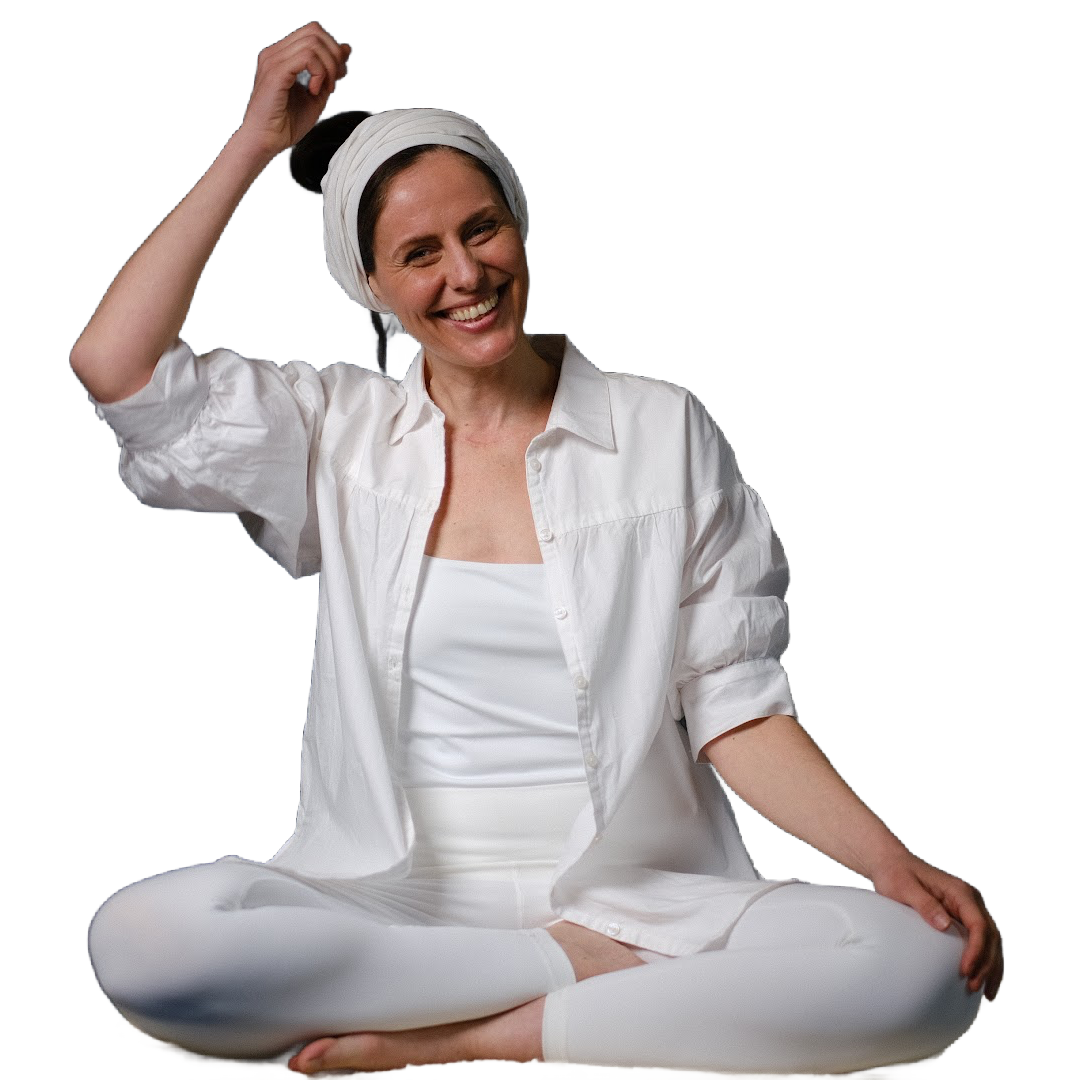 A woman dressed in all white sits cross-legged and smiles, holding one hand above her head, touching her hair. She wears a headscarf, a buttoned shirt, and leggings, with a relaxed and open posture against a plain background.