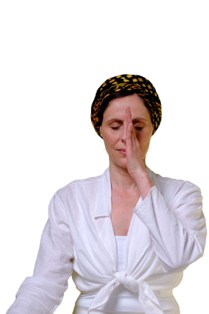 A woman wearing a white top and a patterned headscarf practices alternate nostril breathing, closing one nostril with her finger while keeping her eyes closed. The background is plain white.