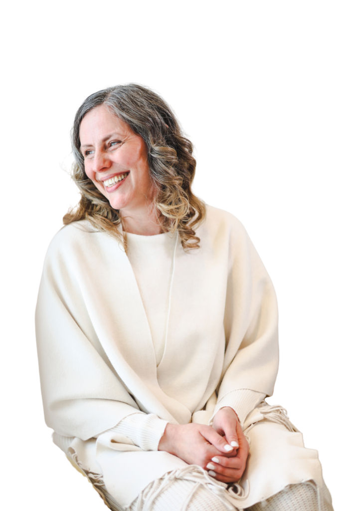 A woman with shoulder-length wavy gray hair, wearing a cream-colored shawl, sits and smiles while looking to her left against a plain white background. Her hands rest in her lap.