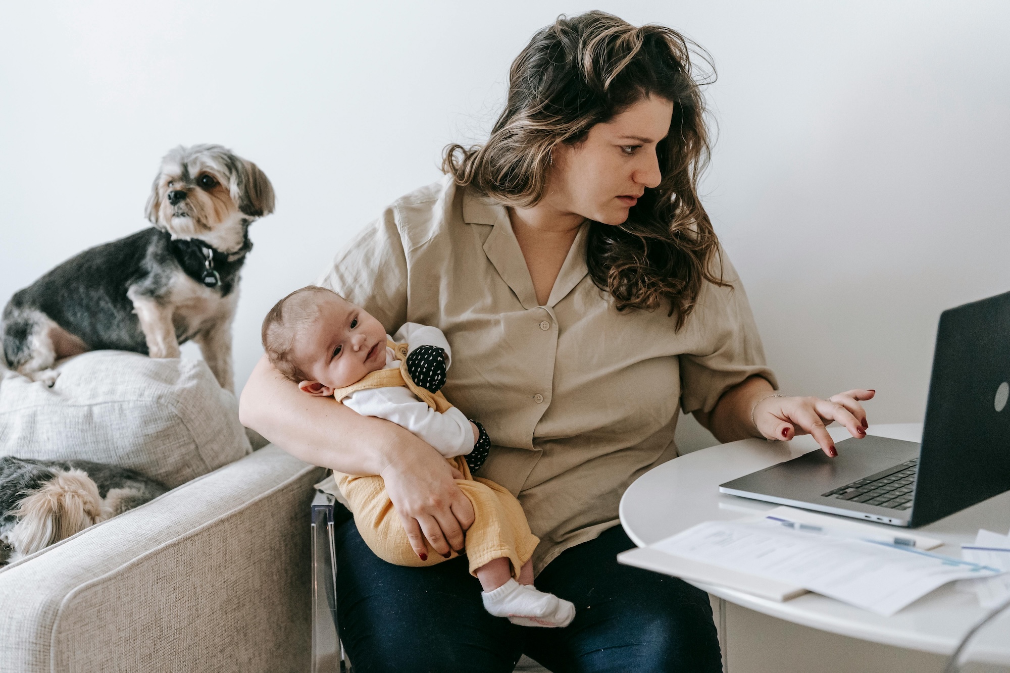 A woman sits at a table working on a laptop while holding a baby on her lap. Papers and a pen are on the table. A small dog sits on the couch behind her, looking to the side.