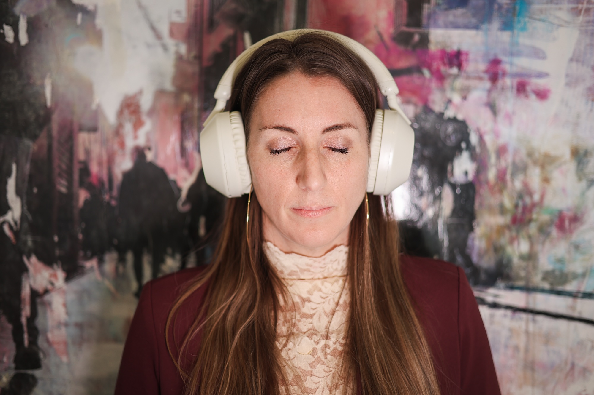 A woman with long brown hair and closed eyes wears large white headphones. She is dressed in a lace blouse and maroon jacket, standing in front of a blurred, abstract background.